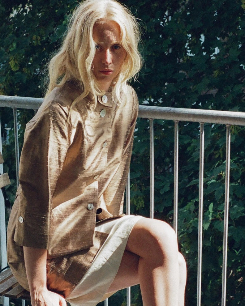 A blonde woman sits on a balcony wearing a loose, light brown linen shirt dress with a cream-colored skirt. The background features a metal railing and lush green foliage.